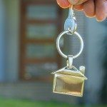 Hand holding a house key in front of a house. The keyring has a house shaped icon on the end and is shiny silver colour. Only the front door is visible in the background. The keyring house icon is dangling down. Close up with shallow focus. Home ownership, new house concept.